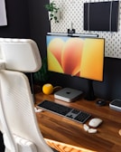 A modern, organized workspace featuring a sleek computer monitor displaying an abstract, colorful wallpaper. On the wooden desk are a keyboard, mouse, and a compact computer unit resembling a Mac Mini. A bright yellow smart speaker and various desk accessories are neatly arranged. A comfortable white office chair is positioned in front of the desk. Behind the monitor, a pegboard with a small plant and a notebook offers storage and decoration options against a dark wall.