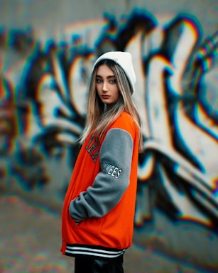 A young individual wearing a bright orange and gray varsity jacket and a white beanie stands in front of a colorful, abstract graffiti wall. The graffiti consists of bold, fluid lines with a mix of blue, orange, and black shades.