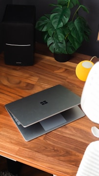 A wooden desk with a partially open laptop displaying the Microsoft logo, a black speaker, a green potted plant with broad leaves, and a small, yellow speaker. There is also a modern office chair partially visible in the foreground.