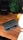 A wooden desk with a partially open laptop displaying the Microsoft logo, a black speaker, a green potted plant with broad leaves, and a small, yellow speaker. There is also a modern office chair partially visible in the foreground.