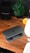 A wooden desk with a partially open laptop displaying the Microsoft logo, a black speaker, a green potted plant with broad leaves, and a small, yellow speaker. There is also a modern office chair partially visible in the foreground.