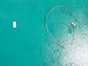 Aerial view of a vast body of turquoise water with a boat on the right deploying a fishing net that creates a circular pattern on the water's surface. A rectangular white platform floats on the left side of the image.