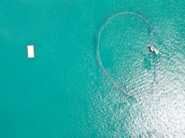 Aerial view of a vast body of turquoise water with a boat on the right deploying a fishing net that creates a circular pattern on the water's surface. A rectangular white platform floats on the left side of the image.