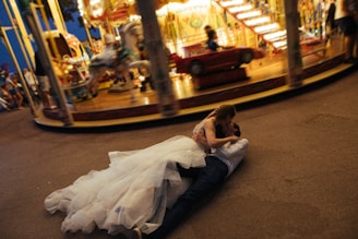 A couple in wedding attire is lying on the ground, embracing each other passionately in front of a carousel. The carousel is in motion, creating a blurred effect of lights and colors in the background. The bride is wearing a voluminous white dress, while the groom is dressed in a shirt and dark trousers. The scene suggests a lively and whimsical atmosphere at a fair.