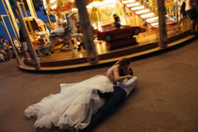 A couple in wedding attire is lying on the ground, embracing each other passionately in front of a carousel. The carousel is in motion, creating a blurred effect of lights and colors in the background. The bride is wearing a voluminous white dress, while the groom is dressed in a shirt and dark trousers. The scene suggests a lively and whimsical atmosphere at a fair.