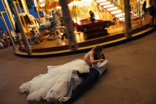 A couple in wedding attire is lying on the ground, embracing each other passionately in front of a carousel. The carousel is in motion, creating a blurred effect of lights and colors in the background. The bride is wearing a voluminous white dress, while the groom is dressed in a shirt and dark trousers. The scene suggests a lively and whimsical atmosphere at a fair.