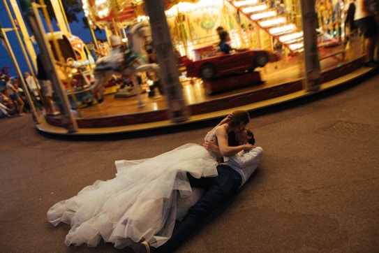 A couple in wedding attire is lying on the ground, embracing each other passionately in front of a carousel. The carousel is in motion, creating a blurred effect of lights and colors in the background. The bride is wearing a voluminous white dress, while the groom is dressed in a shirt and dark trousers. The scene suggests a lively and whimsical atmosphere at a fair.