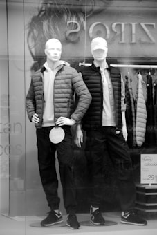 Two mannequins are displayed in a store window, dressed in casual winter clothing. Both mannequins wear puffer jackets over light shirts, paired with dark pants and sneakers. One wears a cap, adding to the casual look. Price tags are visible on the clothing.