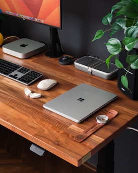A wooden desk with a modern, minimalistic setup including a closed laptop, a keyboard, a mouse, and a monitor. Various small tech gadgets and accessories are neatly arranged on the desk. There is a plant with green leaves adding a touch of nature to the workspace.