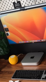 A minimalist desk setup featuring a monitor with an orange and yellow gradient as the background, a Mac mini, and a yellow smart speaker. A keyboard is placed on a wooden desk surface, alongside a green plant.