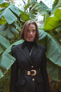A person with short brown hair wearing a black blazer stands in front of lush green banana leaves. The blazer has gold buttons and a decorative belt buckle. The backdrop of dense leaves creates a tropical setting.