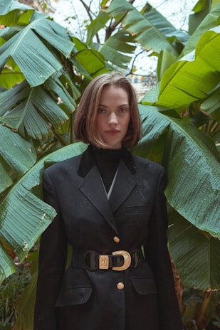 A person with short brown hair wearing a black blazer stands in front of lush green banana leaves. The blazer has gold buttons and a decorative belt buckle. The backdrop of dense leaves creates a tropical setting.