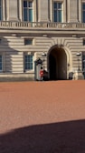 A uniformed guard stands outside a grand historic building with classical columns and arched windows. The guard is wearing a red coat and black hat, positioned near a doorway flanked by two traditional lanterns. The courtyard in front is made of red gravel and is partly covered in shadow.