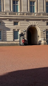 A uniformed guard stands outside a grand historic building with classical columns and arched windows. The guard is wearing a red coat and black hat, positioned near a doorway flanked by two traditional lanterns. The courtyard in front is made of red gravel and is partly covered in shadow.