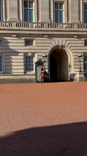 A uniformed guard stands outside a grand historic building with classical columns and arched windows. The guard is wearing a red coat and black hat, positioned near a doorway flanked by two traditional lanterns. The courtyard in front is made of red gravel and is partly covered in shadow.