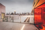 A modern rooftop terrace features a blend of open space and architectural elements, with a panoramic view of a city's skyline. The ground is paved with large tiles, and a glass railing runs along the edge. To the right, a red-tinted glass wall adds a vibrant splash of color. In the distance, illuminated skyscrapers contrast against the overcast sky.