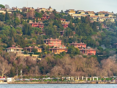 A scenic hillside is lined with a series of residential houses surrounded by lush greenery and trees. The homes are multi-leveled with red roofs, and the landscape slopes down to a waterfront area. People are seen walking near the water, and several cars are parked along the road nearby.