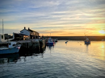 A peaceful harbor scene with a few boats docked by the pier, the water reflects the golden hues of the setting sun, and small buildings line the waterfront. The sky is a blend of soft blues and oranges.