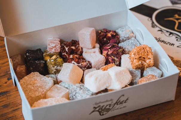 An assortment of colorful Turkish delight neatly arranged in an open white box. The confections come in various shapes, sizes, and textures, featuring coatings of powdered sugar, shredded coconut, and nuts. The box is placed on a wooden table, with a decorative menu or card visible in the background.