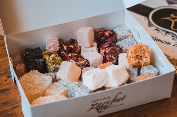 An assortment of colorful Turkish delight neatly arranged in an open white box. The confections come in various shapes, sizes, and textures, featuring coatings of powdered sugar, shredded coconut, and nuts. The box is placed on a wooden table, with a decorative menu or card visible in the background.