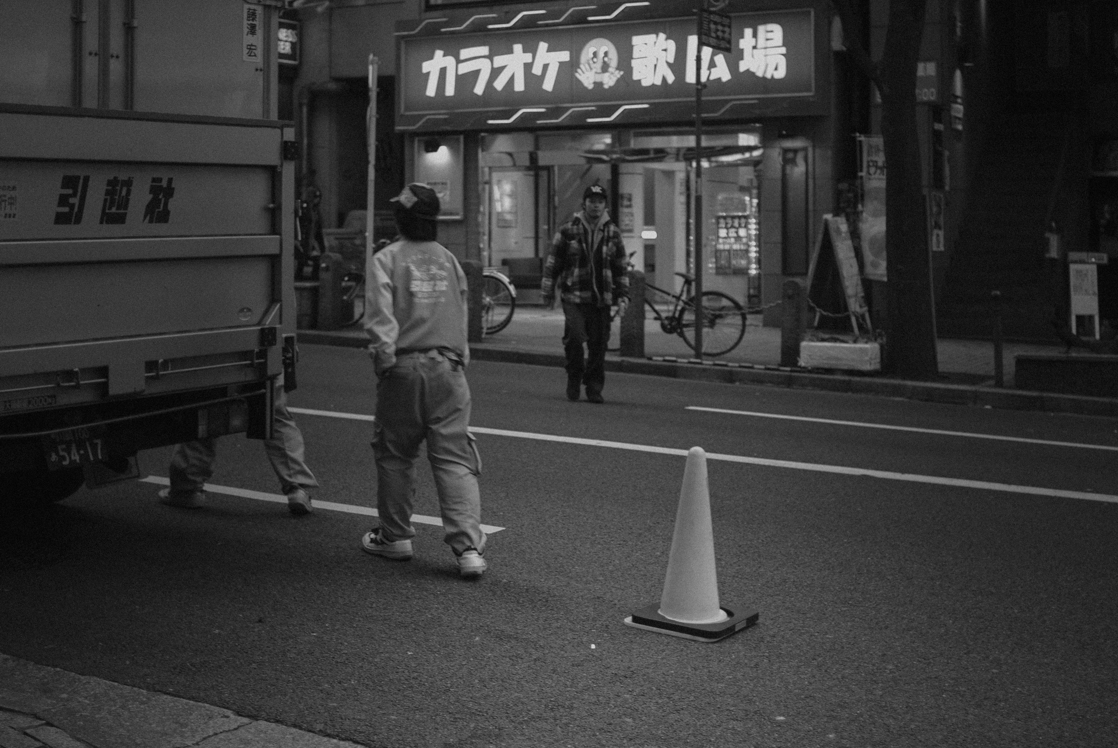 a man standing next to a traffic cone on the side of a road, A small street in Tokyo.