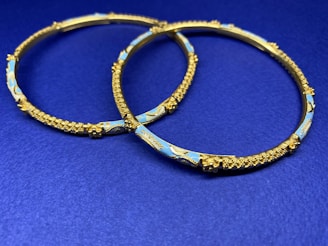 Two ornate gold bangles with intricate detailing and blue enamel decoration are placed on a deep blue surface. The bangles feature textured patterns and delicate craftsmanship.