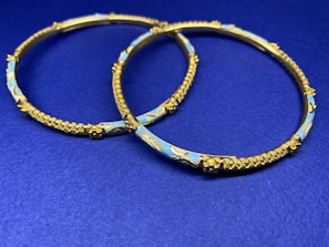 Two ornate gold bangles with intricate detailing and blue enamel decoration are placed on a deep blue surface. The bangles feature textured patterns and delicate craftsmanship.