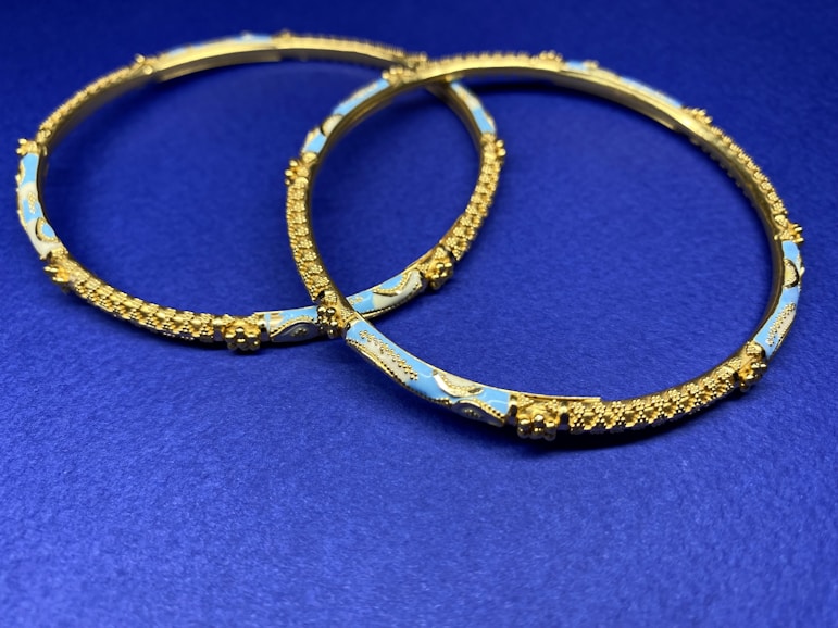 Two ornate gold bangles with intricate detailing and blue enamel decoration are placed on a deep blue surface. The bangles feature textured patterns and delicate craftsmanship.