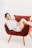 A person is lounging comfortably on a plush, red chair with wooden legs. They are wearing a white shirt and have a relaxed, smiling expression. The background is a plain, light color, enhancing the focus on the person and chair.