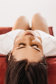 A person with long eyelashes is lying down on their back on a red surface, with their head slightly tilted back. The focus is on their serene face and closed eyes.