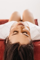 A person with long eyelashes is lying down on their back on a red surface, with their head slightly tilted back. The focus is on their serene face and closed eyes.