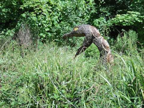 A woven sculpture resembling a creature with an elongated neck and yellow eyes is nestled among dense greenery and tall grass. The sculpture appears to be made of natural materials, blending with the surrounding lush vegetation.