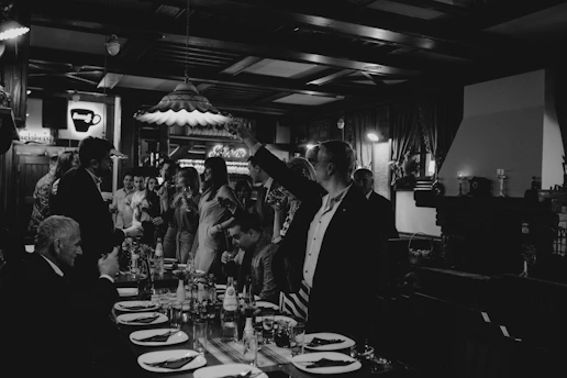 A monochrome image depicting a social gathering in a dimly lit restaurant or bar. People are dressed formally, standing or sitting around a long table set with plates and glasses. The atmosphere suggests a celebration or formal dinner party with attendees holding drinks, engaging in conversation, and exhibiting gestures of toasting.