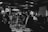 A monochrome image depicting a social gathering in a dimly lit restaurant or bar. People are dressed formally, standing or sitting around a long table set with plates and glasses. The atmosphere suggests a celebration or formal dinner party with attendees holding drinks, engaging in conversation, and exhibiting gestures of toasting.