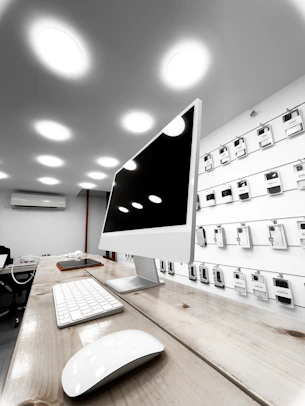 A modern electronics store features a sleek white computer with a large monitor, keyboard, and mouse positioned on a wooden table. The background showcases a wall display of various electronic accessories in white packaging, arranged neatly in rows. The room is illuminated by bright circular ceiling lights, giving a clean and modern ambiance.
