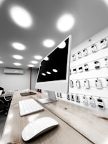A modern electronics store features a sleek white computer with a large monitor, keyboard, and mouse positioned on a wooden table. The background showcases a wall display of various electronic accessories in white packaging, arranged neatly in rows. The room is illuminated by bright circular ceiling lights, giving a clean and modern ambiance.