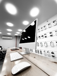 A modern electronics store features a sleek white computer with a large monitor, keyboard, and mouse positioned on a wooden table. The background showcases a wall display of various electronic accessories in white packaging, arranged neatly in rows. The room is illuminated by bright circular ceiling lights, giving a clean and modern ambiance.