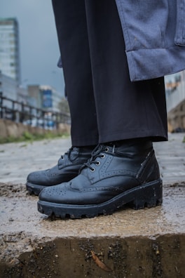 Close-up of a person wearing black leather shoes and standing on a wet concrete step. The shoes have a rugged sole and decorative perforations. The person is also wearing black pants and a blue coat. In the background, there are blurred urban buildings and a cloudy sky, giving a sense of a rainy day.