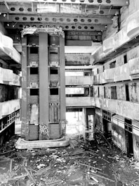 An abandoned multi-story building with exposed concrete, central elevator shafts, and visible graffiti and clutter on the floors. The space appears neglected and in disrepair, with broken beams and debris scattered throughout.