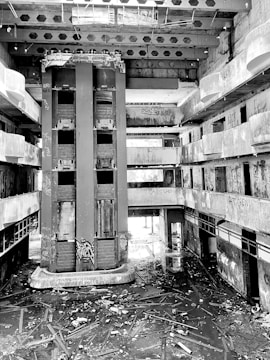 An abandoned multi-story building with exposed concrete, central elevator shafts, and visible graffiti and clutter on the floors. The space appears neglected and in disrepair, with broken beams and debris scattered throughout.