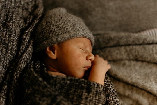 A newborn baby is peacefully sleeping, wrapped in a cozy knitted blanket and wearing a matching knit hat. The texture of the knitted fabric surrounds and envelops the baby, creating a warm and comforting atmosphere.