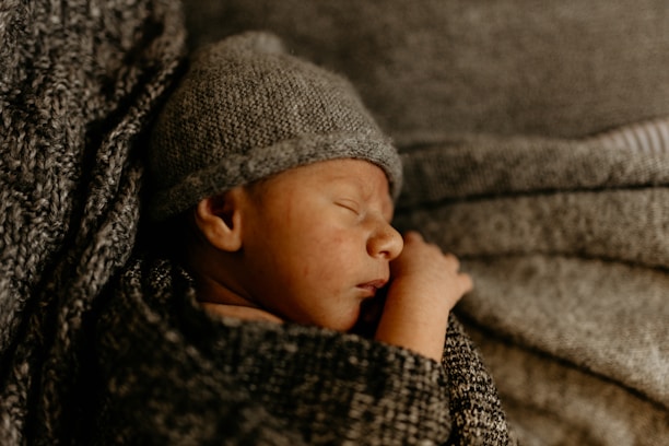 A newborn baby is peacefully sleeping, wrapped in a cozy knitted blanket and wearing a matching knit hat. The texture of the knitted fabric surrounds and envelops the baby, creating a warm and comforting atmosphere.