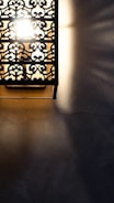 A decorative wall light with intricate metalwork, casting patterned shadows on a textured surface. The light is bright, creating a stark contrast between the illuminated and shadowed areas.