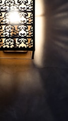 A decorative wall light with intricate metalwork, casting patterned shadows on a textured surface. The light is bright, creating a stark contrast between the illuminated and shadowed areas.