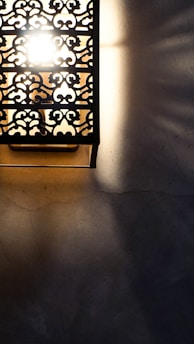 A decorative wall light with intricate metalwork, casting patterned shadows on a textured surface. The light is bright, creating a stark contrast between the illuminated and shadowed areas.