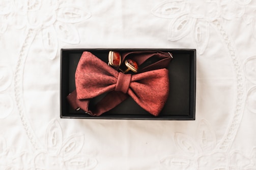 A red bow tie with a subtle pattern is neatly placed in a black rectangular box alongside a pair of cufflinks. The background features a lightly textured white fabric with floral embroidery.