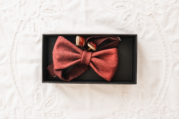 A red bow tie with a subtle pattern is neatly placed in a black rectangular box alongside a pair of cufflinks. The background features a lightly textured white fabric with floral embroidery.