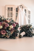 A white wedding dress is hanging on a hanger in the background, draped over a cabinet with glass doors. In the foreground, there are two bridal bouquets featuring a mix of pink, purple, and white roses along with other flowers and greenery, laid out on a white surface.