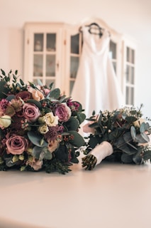A white wedding dress is hanging on a hanger in the background, draped over a cabinet with glass doors. In the foreground, there are two bridal bouquets featuring a mix of pink, purple, and white roses along with other flowers and greenery, laid out on a white surface.