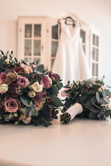 A white wedding dress is hanging on a hanger in the background, draped over a cabinet with glass doors. In the foreground, there are two bridal bouquets featuring a mix of pink, purple, and white roses along with other flowers and greenery, laid out on a white surface.
